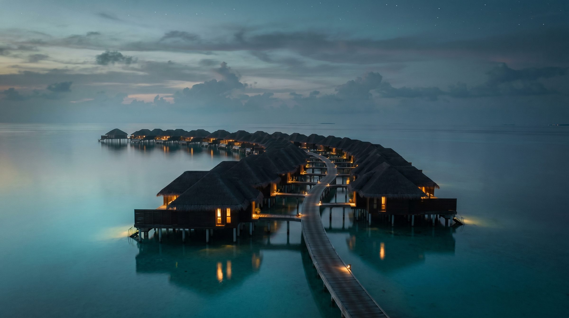 Overwater villas glowing at dusk above a still Indian Ocean lagoon